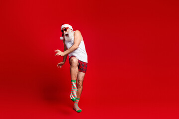 Festive santa lookalike dances in a red studio wearing a santa hat white tank top plaid shorts...