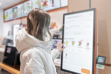 A woman uses a selfservice kiosk in a modern cafe, highlighting tech convenience in dining, A woman in a white jacket chooses drinks and orders food for dessert in a restaurant or pizzeria coffee shop