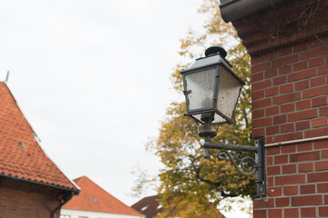 A classic, black square lantern mounted on a decorative, wrought-iron bracket against a red brick wall. The glass panes of the lantern are textured or frosted.