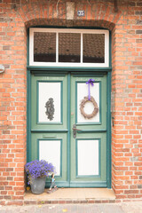 Charming old wooden door painted green and white, framed by red bricks. A lavender wreath and a pot of blooming purple flowers create a welcoming and picturesque rustic entrance.
