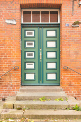 Charming old wooden door painted green and white, framed by red bricks. A lavender wreath and a pot of blooming purple flowers create a welcoming and picturesque rustic entrance.
