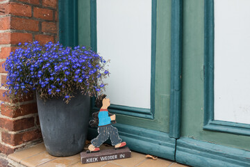 A charming, ground-level view of a doorstep featuring a large, dark gray planter overflowing with vibrant blue flowers (likely Lobelia or Bacopa). Next to the pot, there is a small, whimsical wooden 