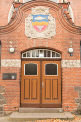 A historic brick building entrance, featuring a double wooden door with a rounded transom window above it. The door is a warm brown color with dark trim. Above the entrance