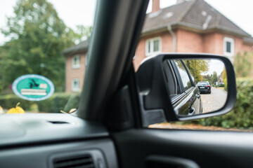 Car side mirror showing a suburban road lined with trees and hedges on an autumn day. A realistic view from inside the vehicle illustrating transportation, safety, and the beauty of seasonal driving.