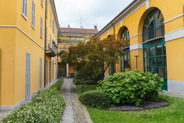 Greenery in the inner yard with pathwalks, historical stone bulding, yellow color, yearly XX century. Monza, Italy.
