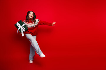 Playful Christmas moment young woman in red sweater spins with large gift against red background...
