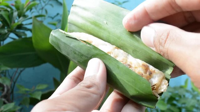 Close-up of two hands unwrapping a piece of traditional cooked food (Lempeng or Serabi) served in a natural green banana leaf wrapper