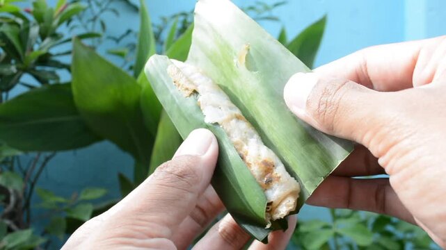 Close-up of two hands unwrapping a piece of traditional cooked food (Lempeng or Serabi) served in a natural green banana leaf wrapper