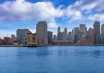 Fototapeta premium Panoramic view of New York City skyline with tall skyscrapers reflecting over calm blue water under a cloudy sky.