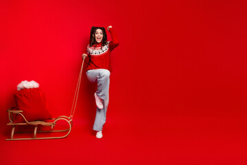 Happy young woman in red Nordic sweater stands by wooden sled with red sack rope red background...