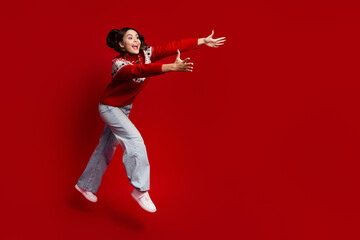 Joyful young woman leaps in a red Christmas sweater against a bold red backdrop capturing festive...