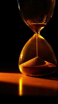 A striking vertical shot captures an illuminated hourglass with golden sand flowing on a reflective surface against a dark background, powerfully conveying time's passage.