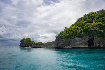 Tropical rocky cliffs with lush green vegetation rising above turquoise sea under dramatic cloudy sky, with a small boat nearby