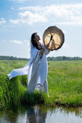 A beautiful female shaman in a trance in the white dress drumming in the natural environment