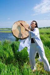 Middle-aged female shaman in the white dress drumming outdoors