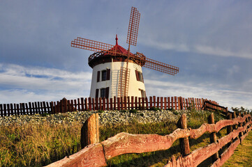 Gostków - Spokojny, kolorowy wiatrak na rustykalnym, drewnianym polu ogrodzonym pod złotym, czystym, błękitnym niebem o zachodzie słońca. © Pol Badger