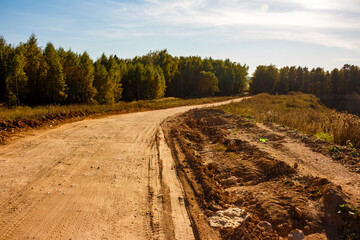 A winding dirt track leads through a rural landscape, flanked by green-golden woods under a clear...