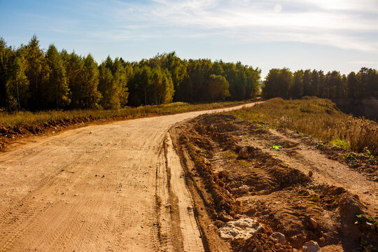 A winding dirt track leads through a rural landscape, flanked by green-golden woods under a clear sky. A rough road by an old quarry on a bright autumn day