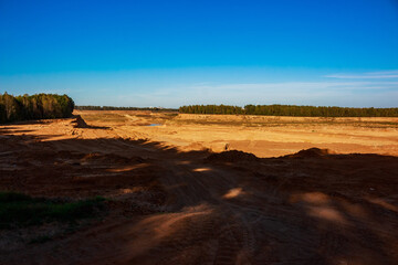 Vast industrial sand quarry under a bright blue sky. Deep tire tracks crisscross the warm, sun-drenched earth, revealing a raw, open-air site