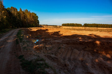 Rugged dirt path through a sprawling sand quarry, bathed in golden light. Autumn foliage lines the distant edges, casting long dramatic shadows