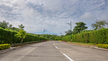 Straight paved road lined with manicured green hedges, streetlights, and trees under a partly cloudy blue sky