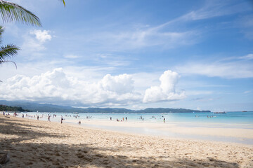 Sunny tropical beach with people swimming and relaxing by turquoise sea and white clouds over distant hills