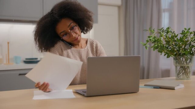 Multitasking African American woman talking mobile phone consider papers managing documents using laptop at home businesswoman busy multitask working remote business paperwork girl female distant job