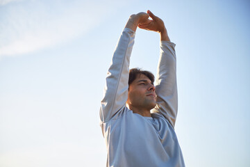 Caucasian young adult man stretching arms overhead outdoors, looking into distance with focused expression, engaging in physical activity against clear sky background