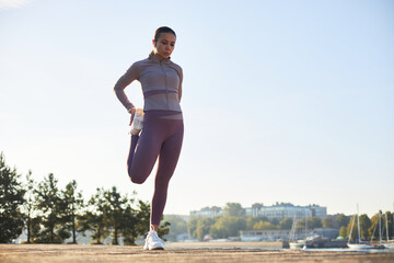 Young adult Caucasian woman stretching leg outdoors on waterfront, preparing for workout, standing...