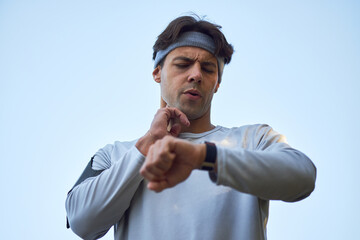 Caucasian young adult man checking pulse on neck while looking at smartwatch during outdoor workout, wearing headband and athletic gear, focused on monitoring heart rate