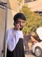 A serious young man with a beard, wearing a casual long-sleeve shirt, stands near a wall talking on a modern smartphone. In the blurred background, a person is seen working on an open car hood