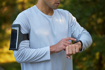 Young adult Caucasian man checking smartwatch while standing outdoors, wearing long sleeve shirt with smartphone armband, focusing on fitness tracking during workout session