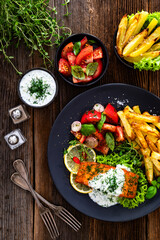 Fried salmon steak with tzatziki sauce, homemade potato fries and tomato salad on wooden table. Top view