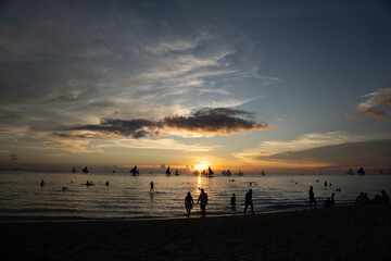 People and sailboats silhouetted against a golden sunset over calm sea at a tropical beach