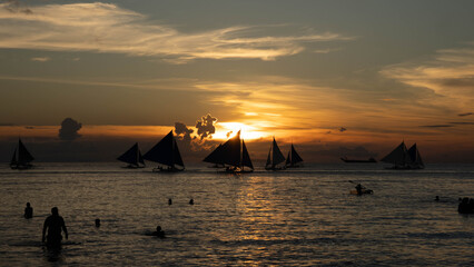 Silhouetted sailboats and people on calm sea during a golden tropical sunset