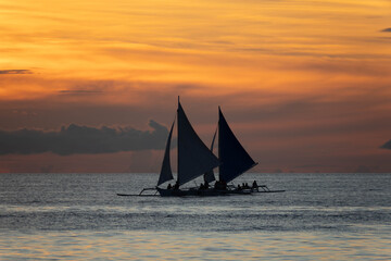 Silhouetted sailboat gliding on calm sea at sunset with vivid orange and golden sky