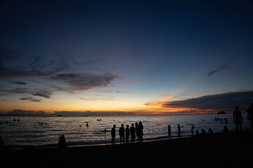 People silhouetted on beach during twilight as the sun sets over calm ocean waves and colorful sky