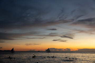 People swimming and sailboats cruising on calm sea during golden sunset with dramatic clouds