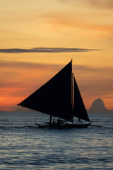 Silhouette of a sailboat gliding over calm sea during golden sunset with orange sky and clouds