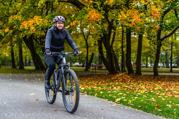Middle aged beautiful woman riding bicycle in city park in autumn. Front view
