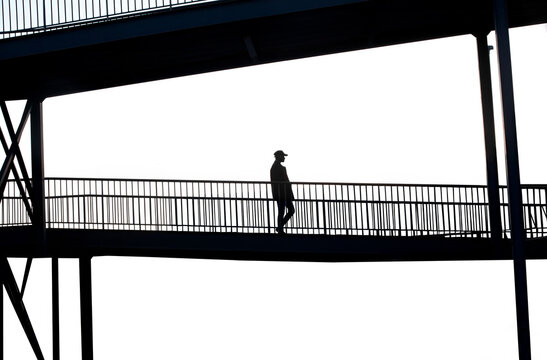 Silhouette of a person walking on a pedestrian bridge. The solitary figure and the stark contrast create a sense of quiet and reflection