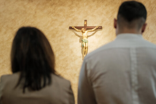 Back view of a religious couple (man and woman) praying and looking at the figure of Christ on the wooden cross (crucifix) on the wall of a church or sacred place. Concept of faith, devotion