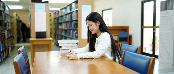Asian young Student doing homework in library of university or college with various book and stationary over the book shelf background, Back to school