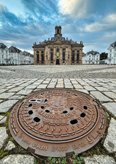 Blick auf einen Kanaldeckel mit Saarbrücker-Wappen und Schriftzug - im Hintergrund die  Ludwigskirche und der Ludwigsplatz