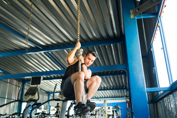 Athletic man climbing rope in gym for fitness workout