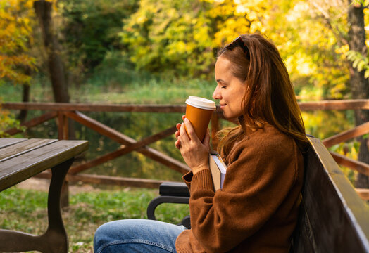 Urban woman enjoying aroma of coffee paper cup while relaxing in autumn park.