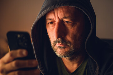 Man wearing black hoodie using smartphone in dark room with warm low key lighting and moody atmosphere.