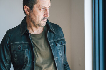 Portrait of confident mature adult man in leather jacket standing by apartment window, looking out at sunny day.