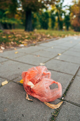 Discarded red plastic shopping bag lying on paved park sidewalk, symbolizing urban waste and environmental pollution, highlighting littering and human impact on nature.
