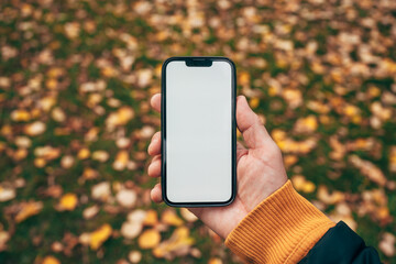 Man holding smartphone with blank white mockup screen in autumn park surrounded by fallen leaves, symbolizing connection and technology outdoors.